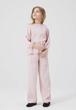 Junges Mädchen in einem hellrosa Langarmoberteil und einer Hose, mit gefalteten Händen stehend und nach oben blickend vor einem schlichten weißen Hintergrund.