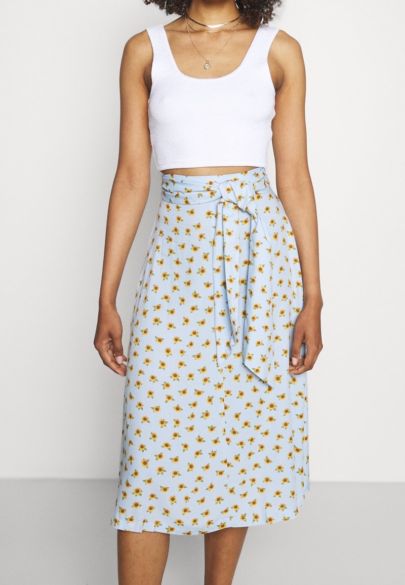 Jupe portefeuille bleu clair avec un lien à la taille, ornée d'un petit imprimé floral jaune. Le tissu semble doux avec une texture lisse.