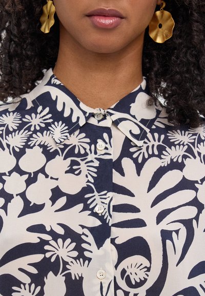 Blusa de estampado floral en colores azul marino y crema, con cierre frontal de botones y un diseño de cuello clásico. Pendientes llamativos en dorado completan el look.