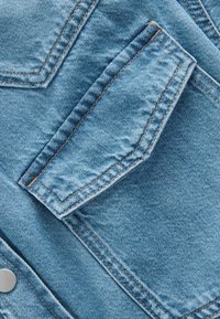 Lichtblau denimstof met een gestructureerd oppervlak, voorzien van een borstzak met schone randen en contrasterende bruine stikseldetails.