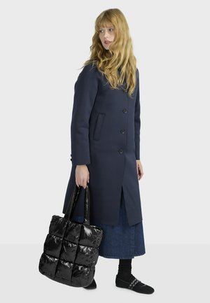 Cappotto lungo blu navy con chiusura a bottoni e tasche, abbinato a una borsa a tracolla trapuntata nera. Il modello indossa una gonna di jeans e calze nere con scarpe.