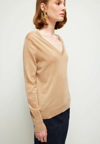 Beige V-Ausschnitt-Pullover aus weichem Material, mit langen Ärmeln und gerippten Bündchen, kombiniert mit einem dunklen Jeansrock.