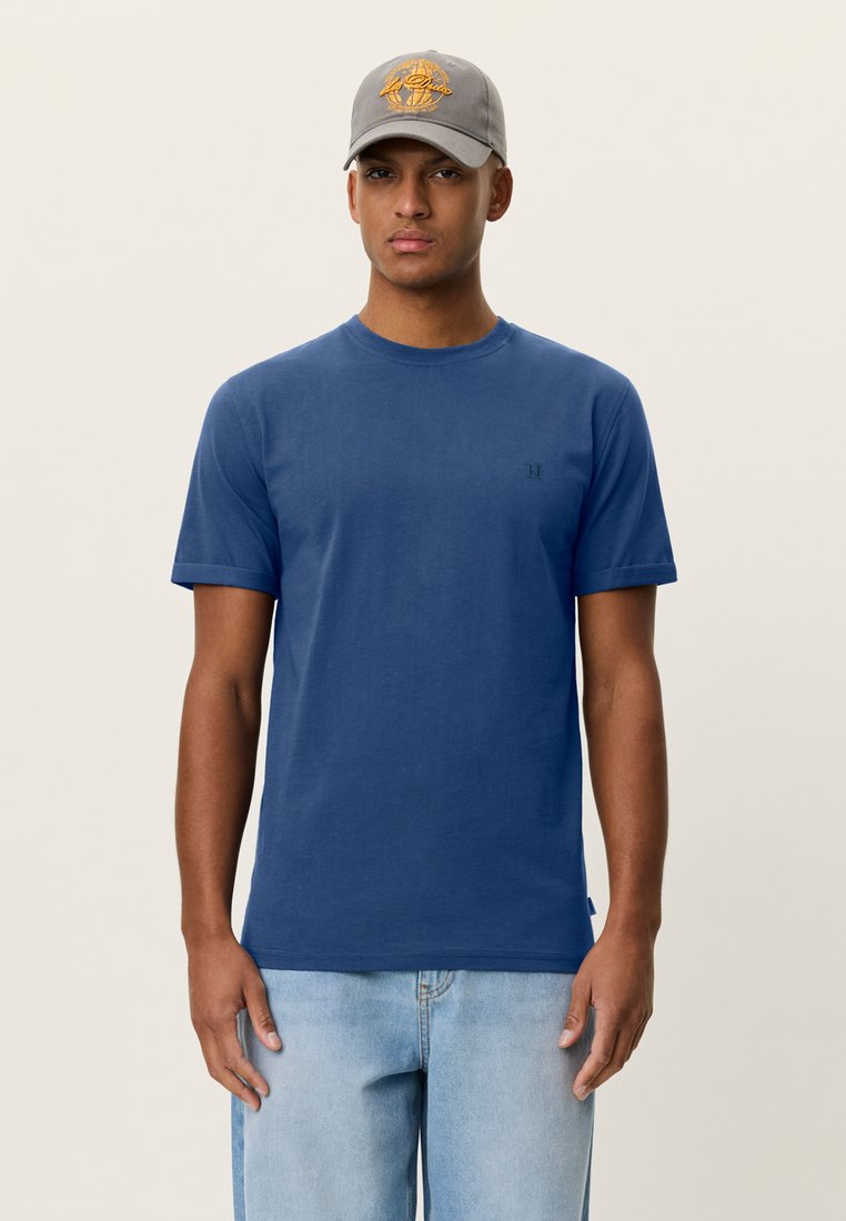 Μπλε βαμβακερό μπλουζάκι με στρογγυλή λαιμόκοψη και κοντά μανίκια, με ένα μικρό πράσινο λογότυπο στο στήθος. Φορεμένο με ανοιχτόχρωμα μπλε τζιν και γκρι καπέλο.