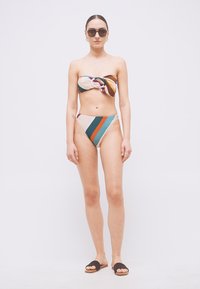 Vrouw draagt een multicolor gestreepte bandeau bikini, donkere zonnebril en zwarte slippers, staand tegen een effen witte achtergrond.