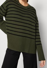 Personne portant un pull en tricot vert olive avec des rayures horizontales noires et un ourlet et des poignets côtelés, associé à un pantalon noir.