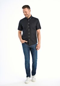 Camicia nera a maniche corte con bottoni e tasca, abbinata a jeans in denim blu e sneakers bianche. Tessuto morbido, vestibilità casual.