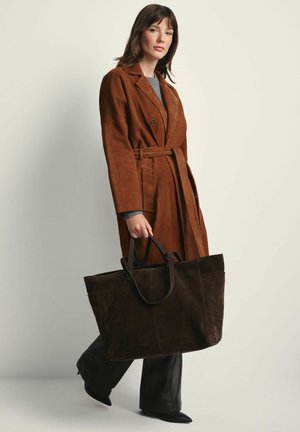 Kobieta w brązowym płaszczu z paskiem i czarnych spodniach, trzymająca dużą ciemnobrązową torbę na ramię, stojąca na jednolitym jasnym tle.