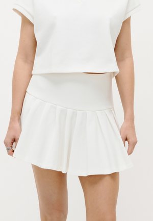 Žena v bílé plisované mini sukni a ladícím krátkém topu, stojící s uvolněnýma rukama podél těla.