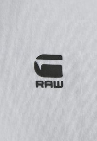 Bílá bavlněná látka s černě potisknutým logem se slovem "RAW" a stylizovaným písmenem "G" nad ním.