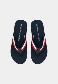 O pereche de flip-flops Tommy Hilfiger de culoare bleumarin, cu curele din material în dungi roșii, albe și bleumarin, pe un fundal alb.