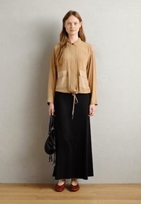 Khaki košeľa na gombíky s dvoma prednými vreckami, skombinovaná s čiernou maxi sukňou a hnedými topánkami na zavretom špičke, držiaca čiernu kabelku.