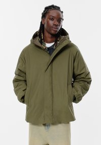 Giubbotto puffer color khaki con cappuccio, dotato di chiusura con zip, tasche laterali e polsini elasticizzati. Texture liscia e design caldo.