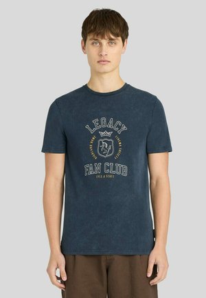 Giovane uomo con capelli corti e castani indossa una maglietta "Legacy Fan Club" blu scuro e pantaloni marroni, in piedi davanti a uno sfondo grigio chiaro uniforme.