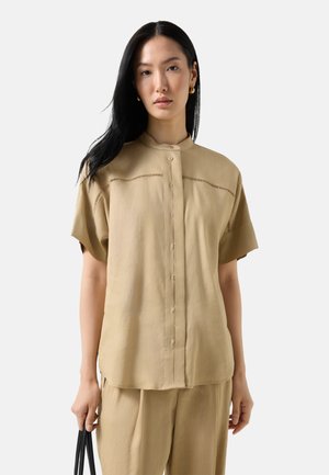 Kvinde med langt sort hår iført beige skjorte med korte ærmer og knapper samt matchende bukser, der holder en sort håndtaske mod en ensfarvet baggrund.