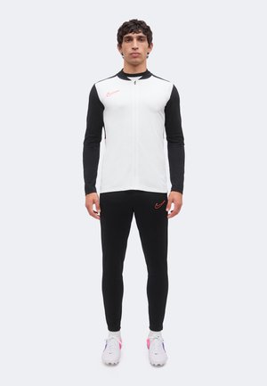 Mężczyzna stoi przodem, ubrany w czarno-białą sportową kurtkę Nike, czarne spodnie z pomarańczowymi logo Nike oraz białe buty sportowe.