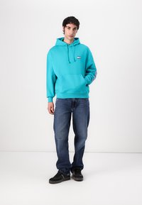 Felpa turchese con tasca frontale e coulisse, con un piccolo logo. Abbinata a jeans azzurri larghi e sneaker nere.