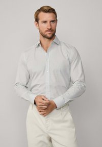 Weißes Hemd mit feinem blauen und schwarzen geometrischen Muster, langärmlig und mit klassischem Kragen. Kombiniert mit hellen Hosen.
