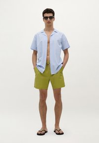 Chemise à manches courtes, rayée bleu clair, déboutonnée au col, assortie à un short vert olive avec poches latérales et cordon de serrage. Tongues noires portées.