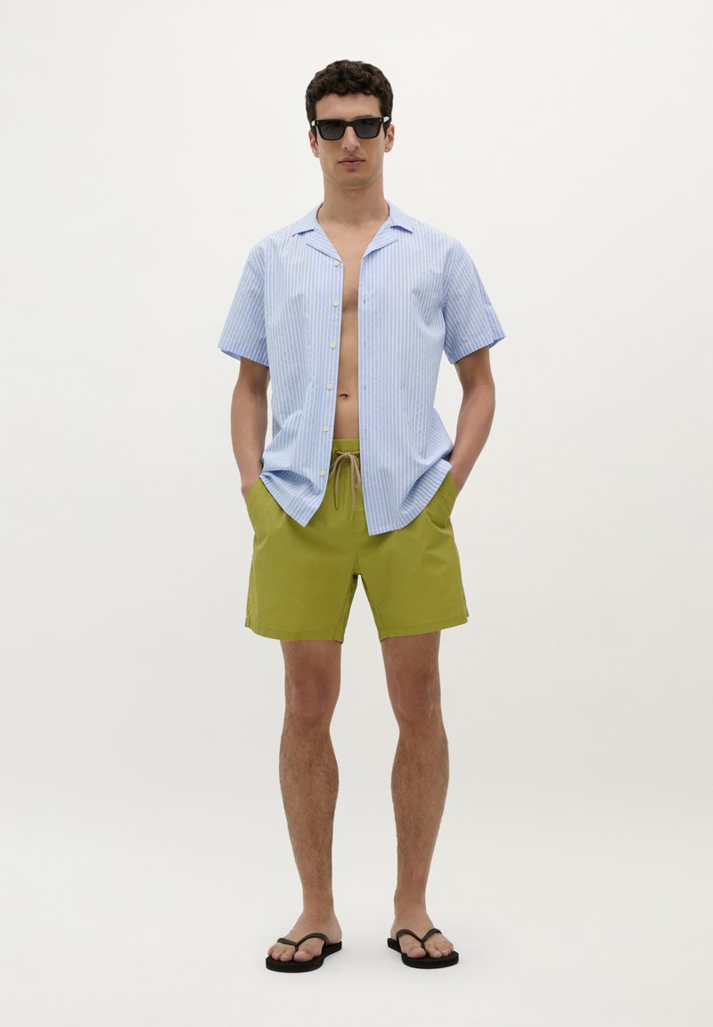 Chemise à manches courtes, rayée bleu clair, déboutonnée au col, assortie à un short vert olive avec poches latérales et cordon de serrage. Tongues noires portées.