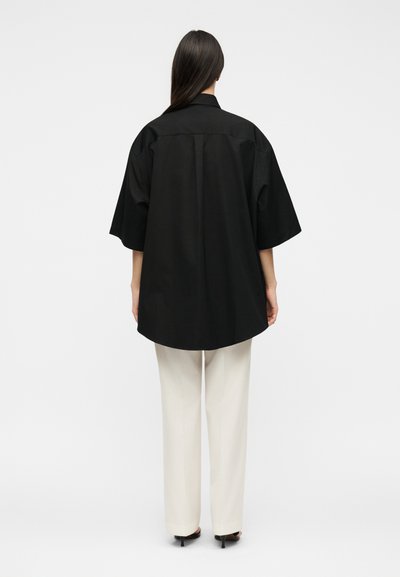 Γυναίκα στέκεται με την πλάτη, φοράει μια μαύρη υπερμεγέθη κοντομάνικη μπλούζα, λευκό παντελόνι και μαύρα σανδάλια με τακούνι σε απλό φόντο.