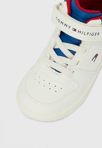 Bílá teniska s modrými akcenty a červeným lemováním, vybavená páskem na suchý zip a perforovanými detaily na špičce. Logo značky na pásku.