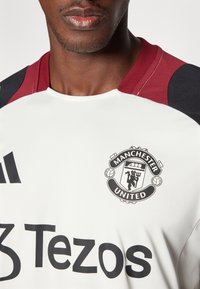 Witte shirt met zwarte en maroon accenten, met het logo van Manchester United en "Tezos" in grote zwarte tekst op de voorkant.