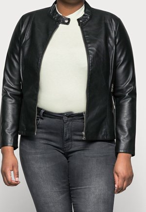 Persoon die een opengezette zwarte leren jas draagt over een witte top, gecombineerd met donkergrijze spijkerbroek, getoond van nek tot heupen tegen een effen achtergrond.