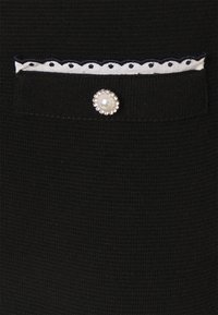 Tissu tricoté noir avec une petite poche dotée d'un galon blanc festonné et d'un bouton rond en perle entouré de petits strass.