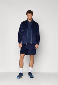 Marineblå kordfløjl jakke med lynlås, parret med en matchende crew-neck trøje og shorts. Afsluttet med blå sportsko og hvide sokker.