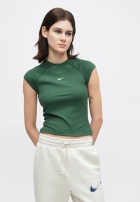 Γυναίκα με σκούρα μαλλιά, φορεμένη με πράσινη αμανική μπλούζα Nike και λευκά παντελόνια φόρμας με κορδόνι και λογότυπο Nike, στέκεται με τα χέρια πίσω από την πλάτη.