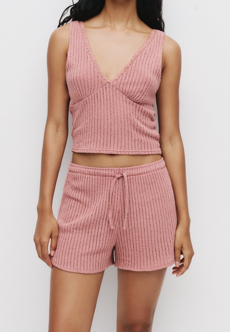 Femme portant un crop top sans manches rose côtelé et un short assorti avec cordon, sur un fond blanc uni.