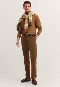 Ein junger Mann steht selbstbewusst in einem enganliegenden braunen Pullover und Cordhosen, umgeben von einem gestreiften Schal und trägt braune Schuhe.