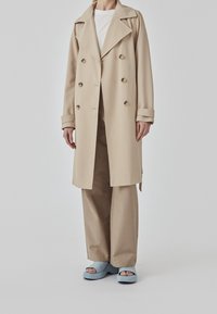Beige double-breasted trenchcoat met grote revers, gecombineerd met beige broek met wijde pijpen en lichtblauwe plateauzandalen. Gladde stoffen textuur.