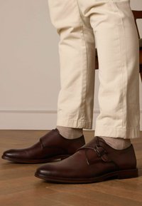 Brown leather dress shoes with a sleek design, rounded toe, and a single buckle accent, paired with light beige trousers and light socks.