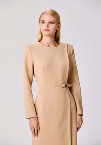 Robe beige à manches longues avec un col rond, un design structuré, un panneau latéral et un détail de ceinture à la taille avec une boucle en argent.