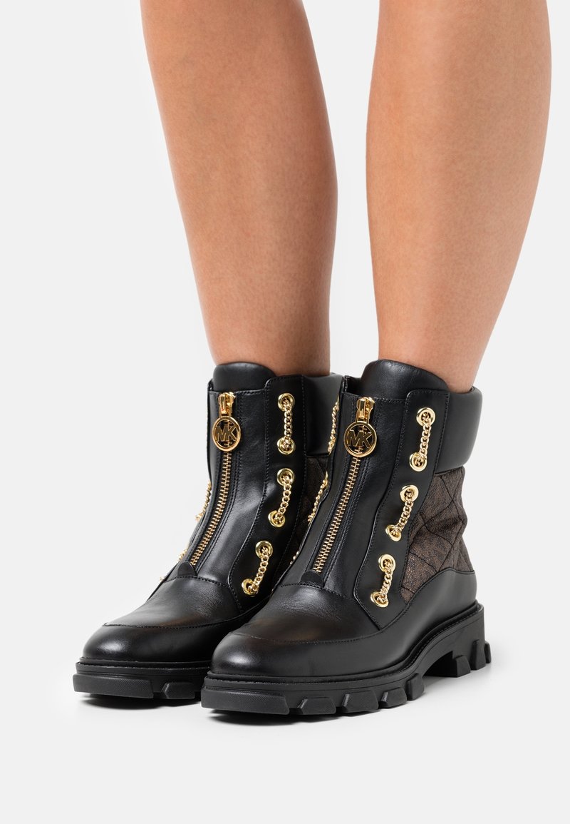 Bottes courtes en cuir noir avec semelles épaisses, fermetures éclair dorées, détails en chaîne et panneaux en tissu matelassé marron, portées sur des jambes nues.