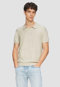 Beige poloshirt met korte mouwen, een v-hals en ribgebreide randen, gemaakt van een textiel met structuur. Gekleed met lichtblauwe spijkerbroek.