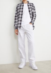 Camicia a quadri bianco e nero, t-shirt bianca, pantaloni bianchi a vestibilità ampia e sneakers bianche. Le trame dei tessuti sono lisce e informali.