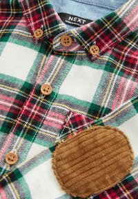 Chemise à carreaux boutonnée avec des carrés verts, rouges, noirs et blancs, boutons en bois et patch marron en velours côtelé au coude.