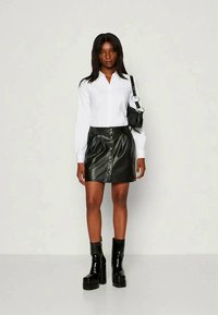 Camisa branca de manga longa com botões, saia mini em couro sintético preto com botões na frente, botas pretas de plataforma, pequena mala preta.