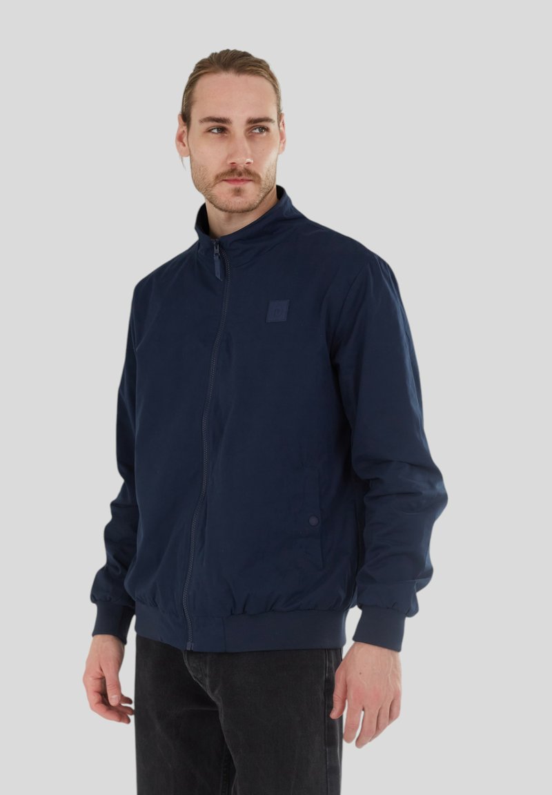 Marinblå zip-up bomberjacka med hög krage, elastiska manschetter och midja. Slät textur och diskret logotyp på bröstet.