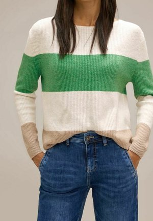Femme portant un pull color-block avec des rayures vertes, blanches et beige, associé à un jean denim bleu, mains dans les poches.