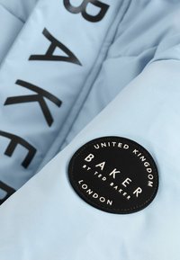 Ljusblå pufferjacka med stora svarta bokstäver som skriver "BAKER." Har en rund svart etikett med vit text som beskriver varumärket och ursprunget.