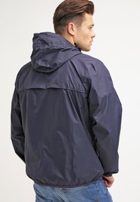 Marineblauwe waterdichte jas met een capuchon, elastische manchetten en een relaxte pasvorm. Soepele textuur en minimale details op de achterkant.