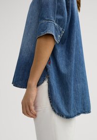 Denim shirt in medium blue, short sleeves, relaxed fit, curved hem, and visible stitching details. Paired with white fabric pants.