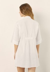 Weißes Leinenkleid mit kurzen Ärmeln, einer elastischen Raffung am Rücken und einer entspannten Passform. Glatte Textur mit einem klaren, minimalistischen Design.