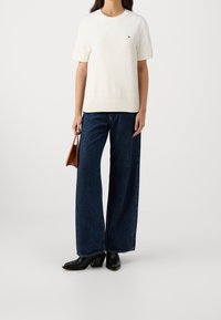 Bílý svetr s krátkým rukávem a kulatým výstřihem, doplněný širokými tmavě modrými džíny. Oblečení je doplněno černými lodičkami na podpatku a hnědou taškou.