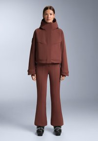 Smeđa izolirana jakna s visokim ovratnikom i dva prednja džepa, uparena s odgovarajućim uskim hlačama s proširenim nogavicama. Tekstura izgleda glatko s ojačanim šavovima.