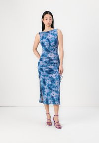 Robe bleue à motifs floraux avec une silhouette ajustée, des détails froncés sur les côtés et des liens ajustables. Associée à des talons strappés violets.
