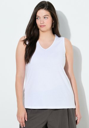 Donna con lunghi capelli scuri che indossa orecchini a cerchio dorati, una maglia bianca senza maniche con scollo a V e pantaloni grigio scuro, in piedi contro un muro bianco.